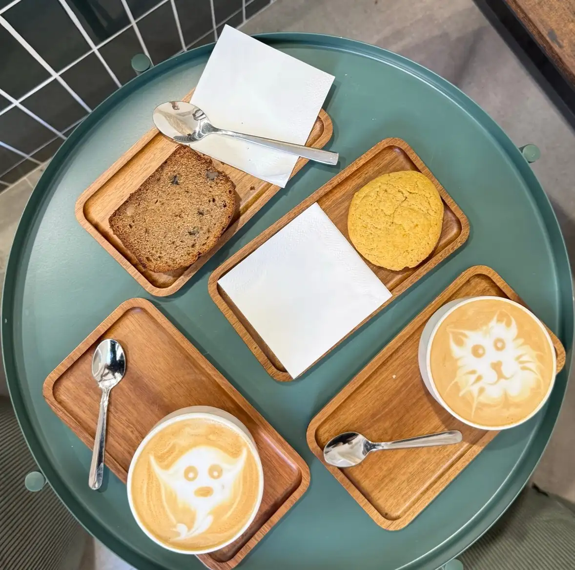 Bandeja redonda con tres cafés con dibujos en la espuma, banana bread y una cookie sobre tablas de madera